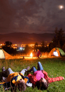 Couple lying under the stars near a campfire and tents in a scenic mountain area – symbolic of relaxation and exploration tied to Good Friday Wellness magic mushroom delivery.