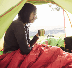 A young woman in a sleeping bag sits inside a tent, holding a metal mug and smiling in the morning light, representing a peaceful moment after a possible mushroom experience outdoors.