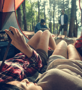 Two people lying inside a tent in the forest, one holding a camera and others relaxing in the background, illustrating a tranquil setting associated with discussions about sleeping on shrooms.