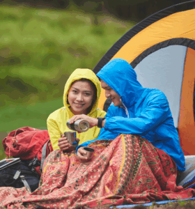 Couple in raincoats sharing a warm drink while camping, with tent and vibrant blanket.