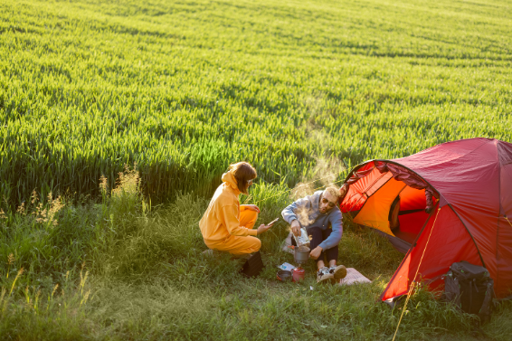 Two women camping in a grassy field next to a red tent, enjoying nature with a hot drink and Good Friday Wellness magic mushroom capsules.