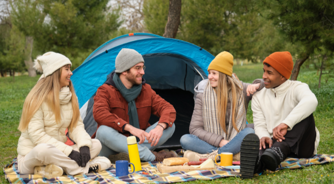 Group of friends smiling and talking outside a tent while camping with mugs and snacks