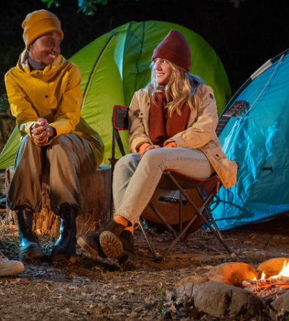 Two campers by a fire at night, smiling and relaxing outside tents.