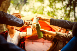 Group of campers toasting drinks in front of a tent in the forest.