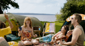 Group of friends laughing and relaxing by tents at a lakeside campsite with a guitar, symbolizing natural wellness and joy