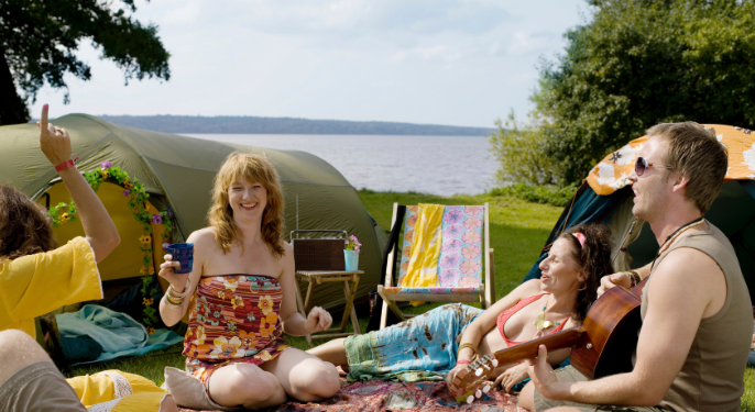 Group of friends laughing and relaxing by tents at a lakeside campsite with a guitar, symbolizing natural wellness and joy