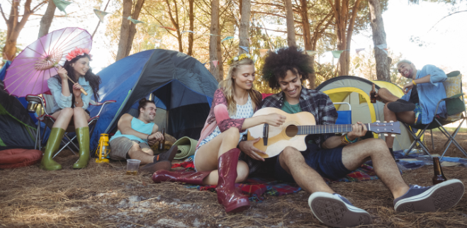 Friends playing guitar, laughing, and relaxing at a lively forest campsite with multiple tents