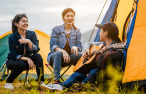 Three women camping with a guitar, smiling and chatting