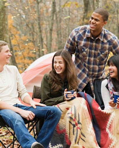 Group of friends camping and laughing in the forest, wrapped in a cozy blanket, symbolizing relaxation and bonding.
