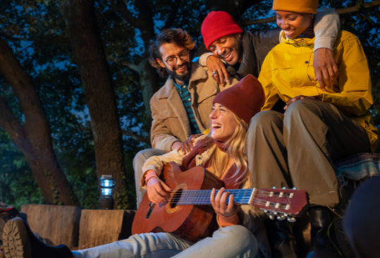 Friends gathered around a campfire laughing as one plays guitar, enjoying nature and connection with Good Friday Wellness magic mushroom capsules.
