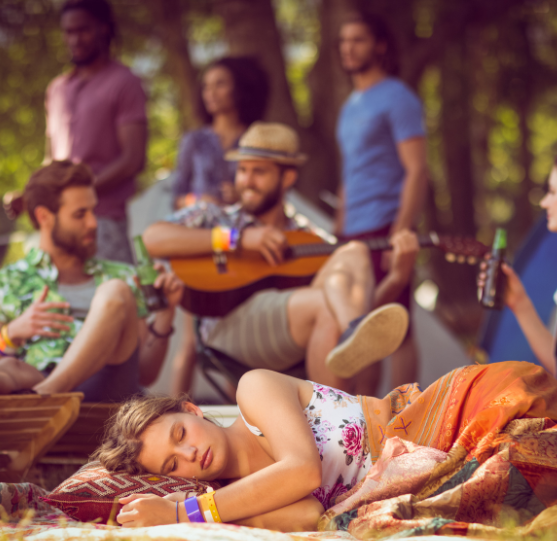 Group of friends at a forest campsite relaxing with music and drinks, one person sleeping peacefully on a blanket