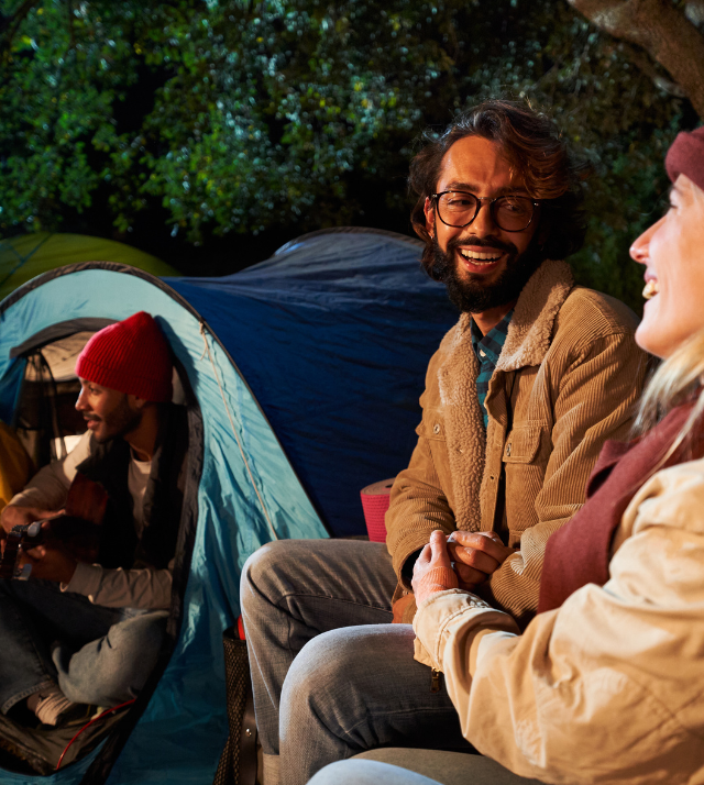 Group of friends sitting at night by tents, smiling and holding hands, with one playing guitar.