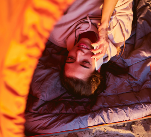 Woman smiling while relaxing in a sleeping bag inside a tent, speaking on the phone