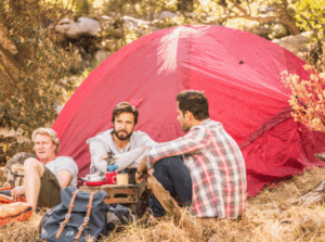 Three men sitting beside a red tent in the forest, engaging in relaxed conversation during a wilderness camping trip.