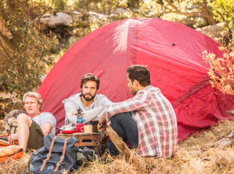 Three men sitting beside a red tent in the forest, engaging in relaxed conversation during a wilderness camping trip.