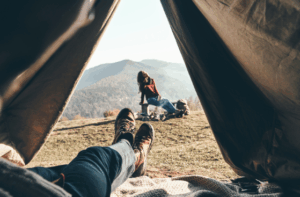 Scenic mountain view from inside a tent, with boots in the foreground and a person relaxing outside—enhanced by Good Friday Wellness magic mushroom delivery.