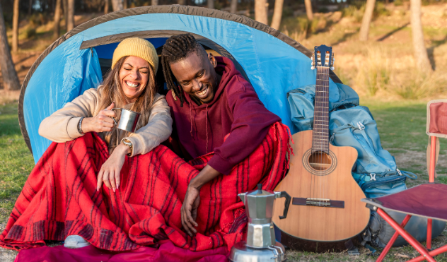 Happy couple camping with coffee and guitar, wrapped in blankets.