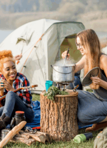 Two women cooking outdoors at a campsite while smiling and playing music.