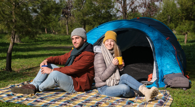 Couple camping in the forest outside a blue tent, enjoying nature with warm drinks.