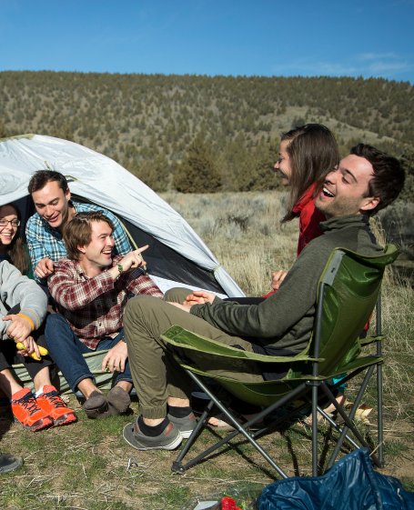 Friends laughing and relaxing at a campsite, enjoying nature with a tent and camping gear, symbolizing the outdoor freedom and wellness experience offered by Good Friday Wellness magic mushroom