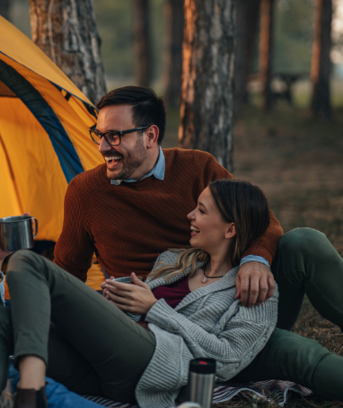 Couple sitting outside a tent with mugs, smiling and enjoying a peaceful forest camping moment